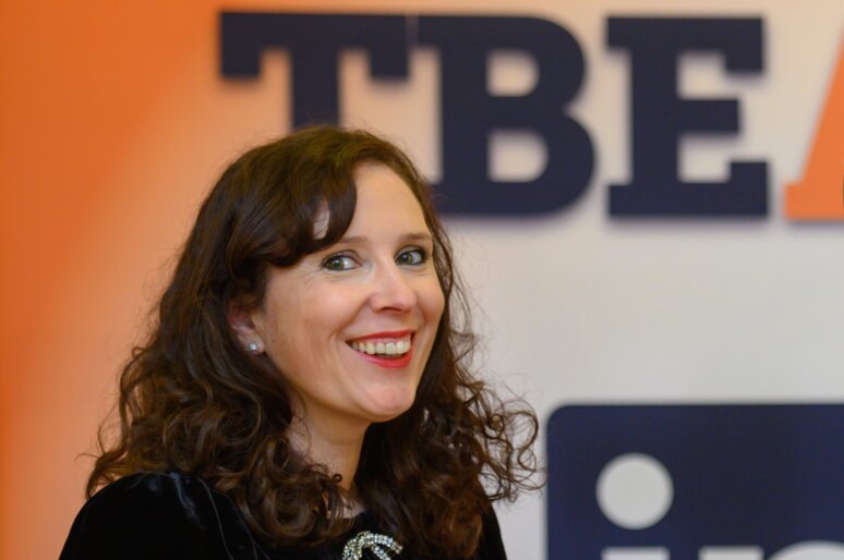 Anita Jaynes, Founder of The Business Exchange South West, smiling at a professional event with the TBE logo visible in the background. She is wearing a black outfit with a decorative brooch.