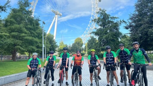A group of eight cyclists in matching Bristol Airport charity cycling jerseys pose with their bikes in front of the London Eye on a sunny day