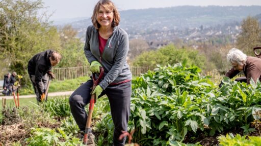 A person smiles while holding a shovel in a lush garden at Bath City Farm, surrounded by thriving plants. They wear a dark jacket, red shirt, gloves and boots. In the background, two others tend to the greenery, with rolling hills and a town visible under a partly cloudy sky—capturing a moment of teamwork, sustainability and connection to nature.
