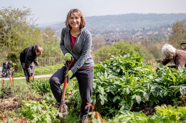 A person smiles while holding a shovel in a lush garden at Bath City Farm, surrounded by thriving plants. They wear a dark jacket, red shirt, gloves and boots. In the background, two others tend to the greenery, with rolling hills and a town visible under a partly cloudy sky—capturing a moment of teamwork, sustainability and connection to nature.