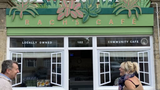 Exterior of Rach’s Café, a locally owned community café with green signage decorated with illustrated plants. The café owners, Andy and Rach, are smiling at each other outside near café tables.