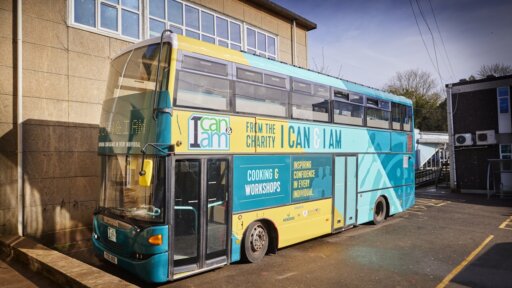 A turquoise and yellow double-decker bus parked beside a building, branded with the charity name ‘I Can & I Am’. The bus features text promoting cooking workshops and inspiring confidence, with large windows on the upper deck.