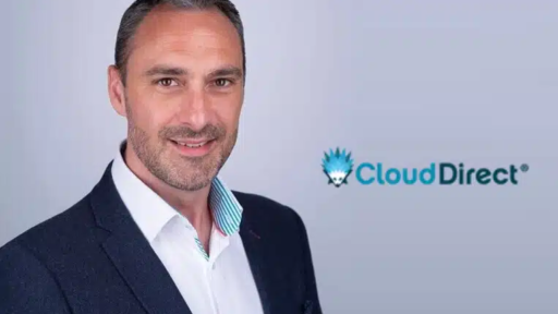 Leighton Searle wearing a dark blazer over a white collared shirt with a light blue and pink striped inner lining, standing against a light grey background. To the right, the Cloud Direct logo is visible, featuring a blue stylized cloud icon with text reading “CloudDirect®” in blue and black.