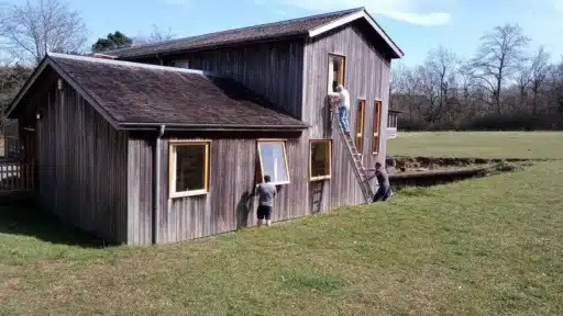 Three people working on a wooden-clad building in a grassy outdoor area under a clear blue sky. One person is on a ladder cleaning or repairing an upper window, another is holding the ladder for support, and a third person is working on a lower window. The building has multiple rectangular windows with wooden frames and a sloped roof, surrounded by open green space and trees in the background.