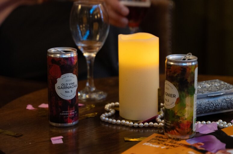 A warm, intimate tabletop scene featuring two Canned Wine Group cans beside a glowing candle, with a wine glass, a string of pearls, confetti and an ornate silver box adding to the festive atmosphere.