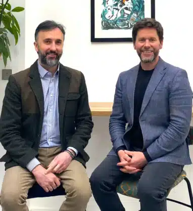 Duality founders Alberto Politit and Anthony Laing seated side by side on stools in a modern office setting, wearing smart‑casual business clothing, with framed artwork on the wall behind them.