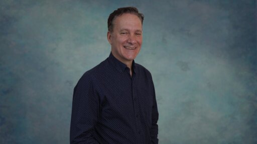 A portrait-style image of Roland Storti standing against a mottled blue and teal studio backdrop. They are wearing a dark long‑sleeved shirt with a subtle dotted pattern, with one hand in their pocket and their body angled slightly to the side.