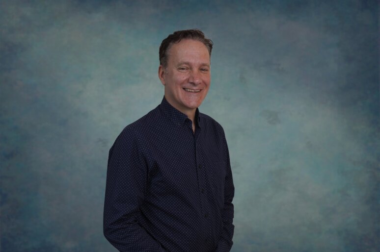 A portrait-style image of Roland Storti standing against a mottled blue and teal studio backdrop. They are wearing a dark long‑sleeved shirt with a subtle dotted pattern, with one hand in their pocket and their body angled slightly to the side.
