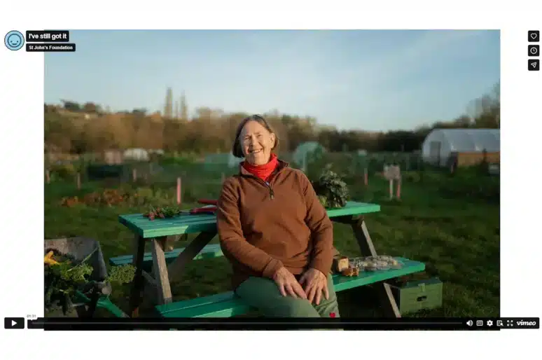 A person sits on a picnic bench in an allotment or community garden. They are wearing a brown fleece, red neck scarf and green trousers. Gardening tools, freshly picked vegetables and small pots are placed on the bench and beside a wheelbarrow nearby. In the background are greenhouses, raised beds and trees under a clear sky. The scene is a video still from a St John’s Foundation project.