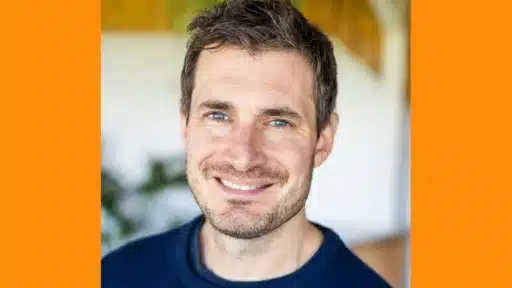Steve Jeffreys wearing a dark navy blue crew-neck sweater is shown from the shoulders up. The background is softly blurred, featuring green foliage and warm-toned wooden elements, suggesting an indoor setting with natural light. The image has orange borders on both sides.