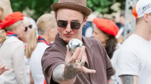 A person wearing a brown hat and short-sleeved shirt holds a shiny metal boules ball toward the camera, with a crowd of people in colourful clothing gathered behind them at an outdoor event.