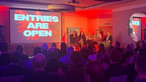 A large audience sits facing a stage lit in red and purple. A big screen displays the words “ENTRIES ARE OPEN” in bold white text over a red splash graphic. Four presenters are seated on stage, with banners for the event behind them. Bath Dragons 2026.