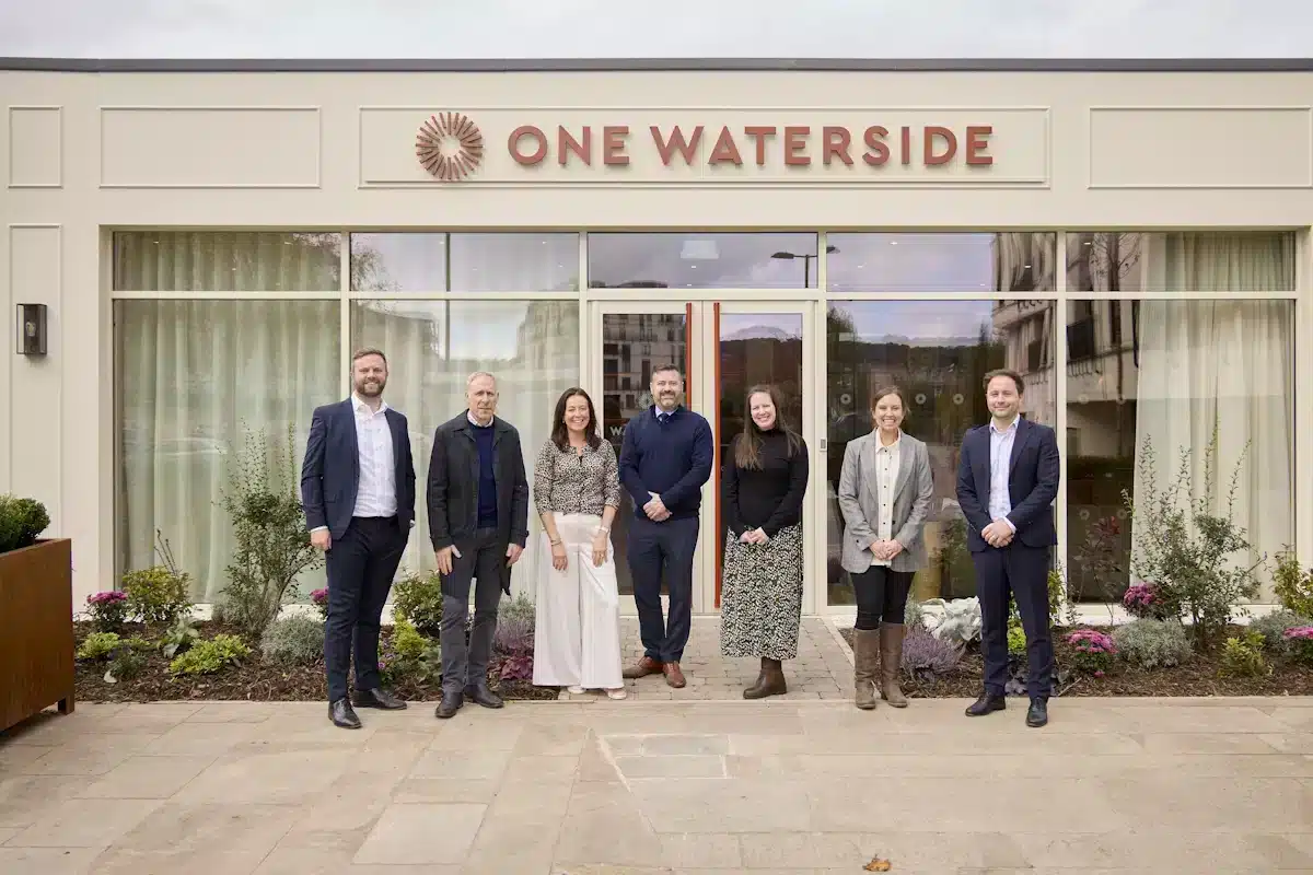A group of seven people standing outside the One Waterside building, positioned in front of landscaped planting and the entrance, with the building’s signage visible above them.