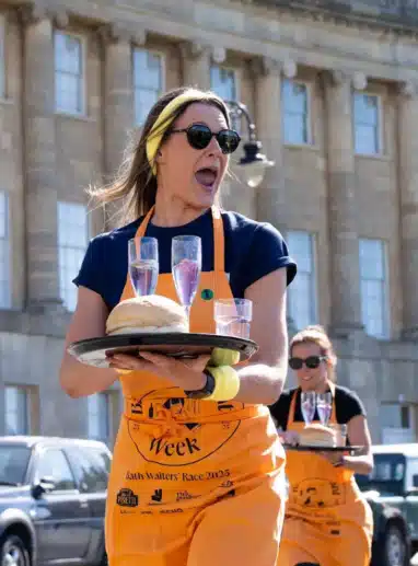 A participant in an orange apron carries a tray holding glasses of water and a bread roll during the Bath Waiters Race. She walks in front of the Royal Crescent on a sunny day, with another competitor following behind and cars parked along the street.