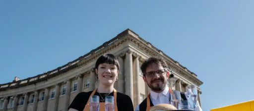 Two Bath Waiters' Race participants wearing aprons carry trays holding bread rolls and glasses of water during an outdoor waiter race event. They stand in front of the Royal Crescent in Bath, with a crowd gathered in the background under a clear blue sky.