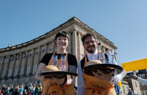 Two Bath Waiters' Race participants wearing aprons carry trays holding bread rolls and glasses of water during an outdoor waiter race event. They stand in front of the Royal Crescent in Bath, with a crowd gathered in the background under a clear blue sky.