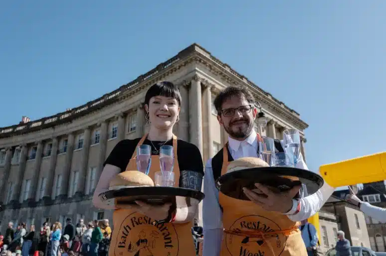 Two Bath Waiters' Race participants wearing aprons carry trays holding bread rolls and glasses of water during an outdoor waiter race event. They stand in front of the Royal Crescent in Bath, with a crowd gathered in the background under a clear blue sky.