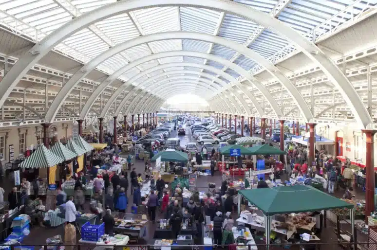A busy indoor market at Green Park Station with a high arched glass roof, filled with stalls selling food, crafts and various goods. Green and white striped canopies and green gazebos are scattered throughout the space, while shoppers walk between the stalls. The hall is bright with natural light, and parked cars are visible toward the far end of the building.