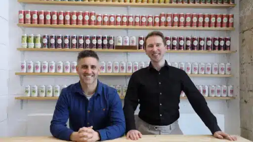 Simon Rollings and Ben Franks positioned behind a wooden counter in a shop or tasting room. Simon is seated and wearing a blue shirt, while Ben stands beside him wearing a black shirt. Behind them, wall‑mounted shelves are filled with neatly arranged cans in rows of different colours and flavours. The setting has a bright, modern, industrial feel with stone and white walls.