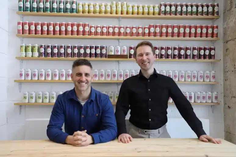 Simon Rollings and Ben Franks positioned behind a wooden counter in a shop or tasting room. Simon is seated and wearing a blue shirt, while Ben stands beside him wearing a black shirt. Behind them, wall‑mounted shelves are filled with neatly arranged cans in rows of different colours and flavours. The setting has a bright, modern, industrial feel with stone and white walls.