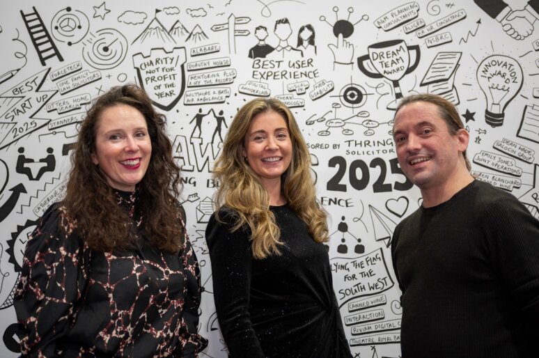 Anita, Anna and Ben from The Business Exchange stand in front of a white illustrated backdrop filled with hand‑drawn icons, awards, and motivational phrases. The person on the left wears a patterned black and pink top, the person in the centre wears a black dress with subtle shimmer, and the person on the right wears a dark long‑sleeve shirt. The backdrop features sketches such as “Best Start Up 2023,” “Best User Experience,” “Charity Not for Profit of the Year,” and various doodles related to innovation, collaboration, and awards.