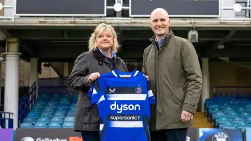 Two people standing in a rugby stadium holding up a blue, black, and white Bath Rugby shirt with “Dyson Supersonic” printed on the front. They are positioned in front of empty blue seats and stadium signage.