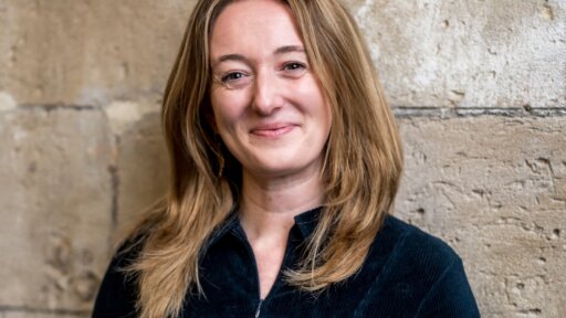 A portrait-style photograph of Becky Somerset standing against a textured stone wall. She has shoulder-length hair and is wearing a dark zip-up top.