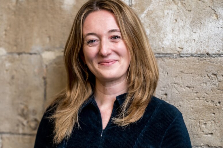 A portrait-style photograph of Becky Somerset standing against a textured stone wall. She has shoulder-length hair and is wearing a dark zip-up top.