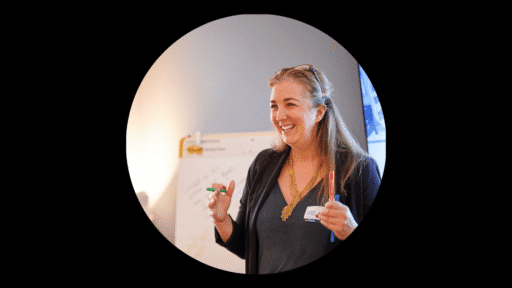 Claire Jonik standing indoors giving a presentation, holding two markers and gesturing with both hands. In the background there is a flip chart with notes and a lit workspace.
