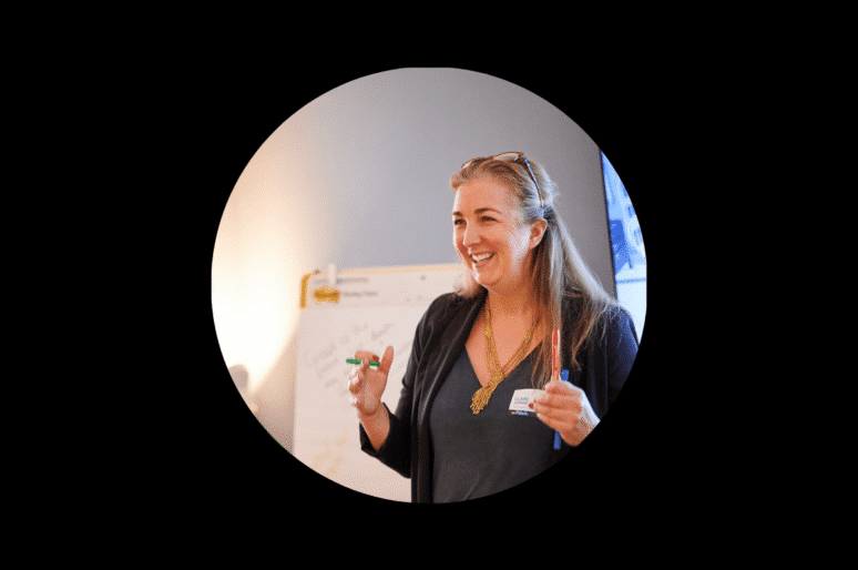 Claire Jonik standing indoors giving a presentation, holding two markers and gesturing with both hands. In the background there is a flip chart with notes and a lit workspace.