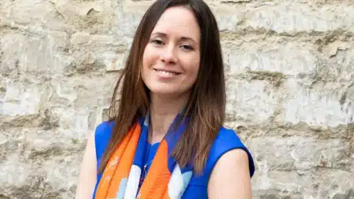Emma Davis ith long brown hair is standing in front of a light‑coloured stone wall, wearing a bright blue sleeveless top and an orange, white, and blue patterned scarf. The image is taken outdoors with natural lighting.