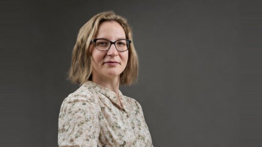 Judith Coghlan sits at a table against a plain dark-grey backdrop, wearing a light-coloured, floral-patterned blouse. A glass of water is placed on the table in the lower right corner of the image.