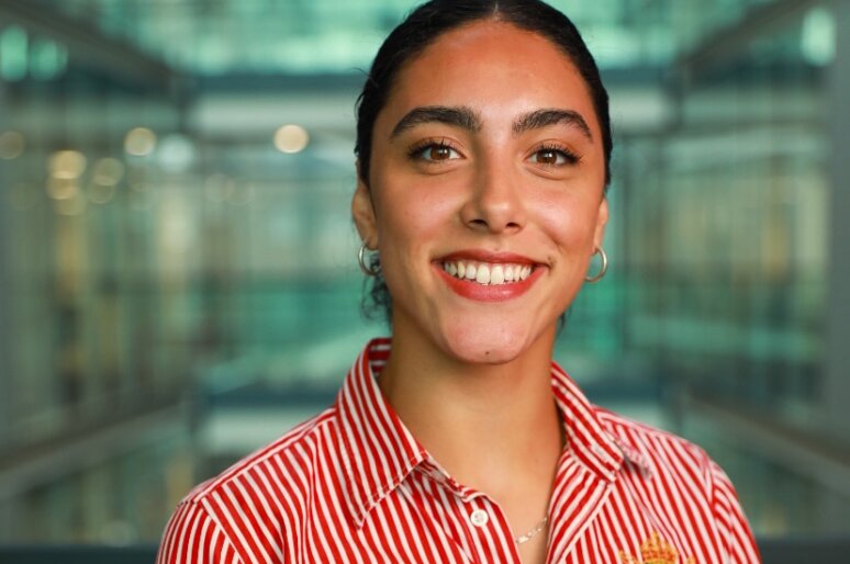 Lauren Bennett earing a red and white striped shirt with an embroidered crest stands indoors in a modern glass building. Soft, natural light fills the background, which features multiple levels, glass panels and metal railings. She is also wearing small hoop earrings and a delicate necklace.
