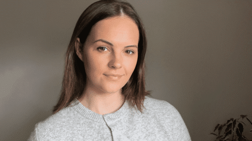 Megan Chiplen wearing a light grey, buttoned knit sweater is positioned indoors against a plain, softly lit wall. She has straight, shoulder‑length brown hair, and a small leafy plant is visible in the lower right corner of the scene.