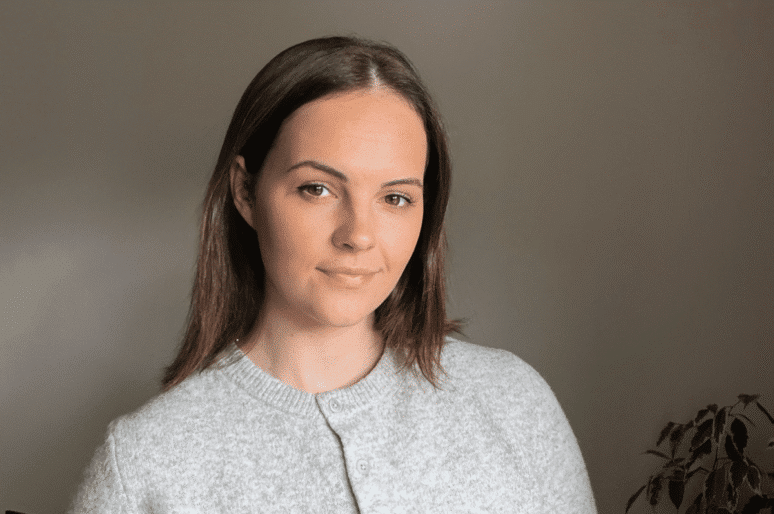 Megan Chiplen wearing a light grey, buttoned knit sweater is positioned indoors against a plain, softly lit wall. She has straight, shoulder‑length brown hair, and a small leafy plant is visible in the lower right corner of the scene.