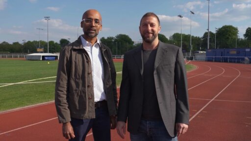 Roy and Dario from ForceTeck stand side by side on an outdoor running track. Both are wearing jackets and trousers, and a sports field with green grass is visible behind them. Floodlights, fencing, and various track‑side structures appear in the background under a partly cloudy sky.