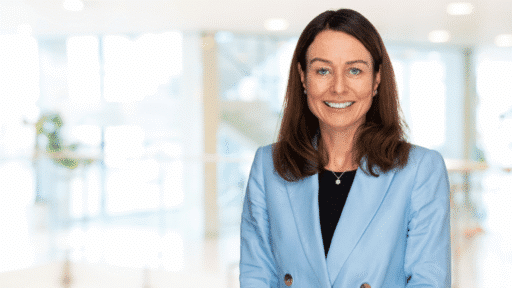 Sarah Loveless wearing a light blue blazer and black top is photographed indoors in a bright, modern office setting with soft, out‑of‑focus background lighting and glass features.