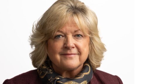 Sophie Hall with light-coloured, shoulder‑length hair is pictured against a plain white background. The person is wearing a textured burgundy jacket and a dark patterned scarf featuring a design with gold tones.