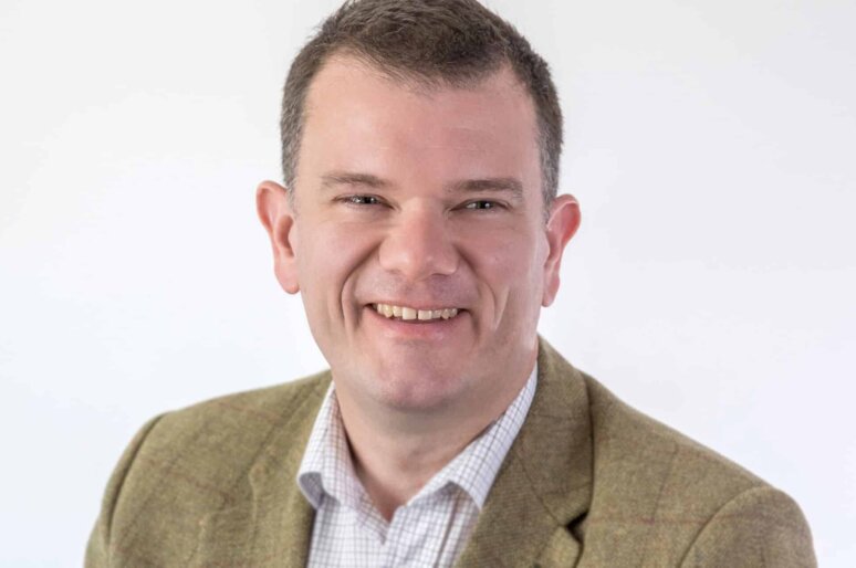 Professional headshot of Harvey Davies wearing olive coloured jacket and white shirt