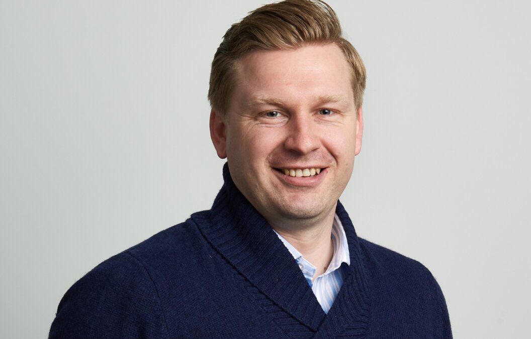 Ed Tellwright smiling and wearing a dark blue sweater with a small red embroidered logo and a light blue collared shirt underneath, posed against a plain light gray background in a clean, professional portrait style.